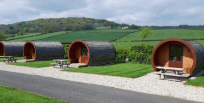 A row of barrel-shaped camping pods on grass with picnic benches and hills behind