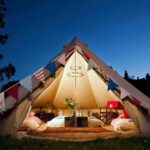 A canvas bell tent glowing from within at dusk, its doors tied open to show floor cushions and lanterns, strung with bunting and a ring of fairy lights