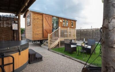 Spinney View shepherd's hut lit up at dusk, its stable door open, with the hot tub, bistro set and swing fire pit on the terrace in front
