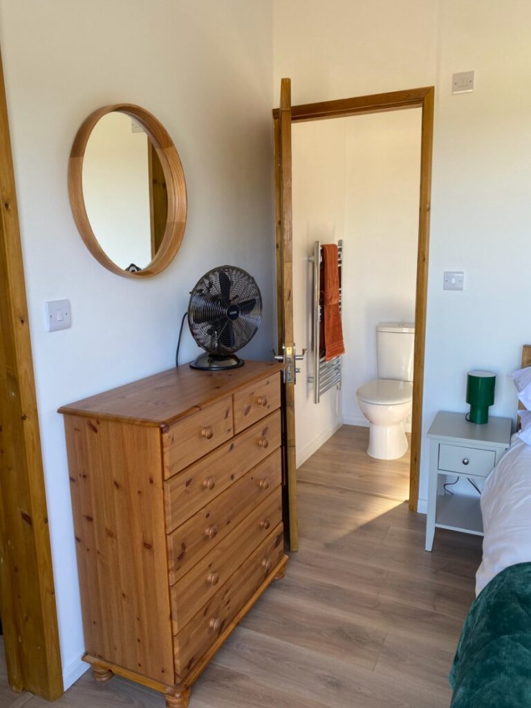 Cabin bedroom corner with a pine chest of drawers, a round mirror and the en-suite door