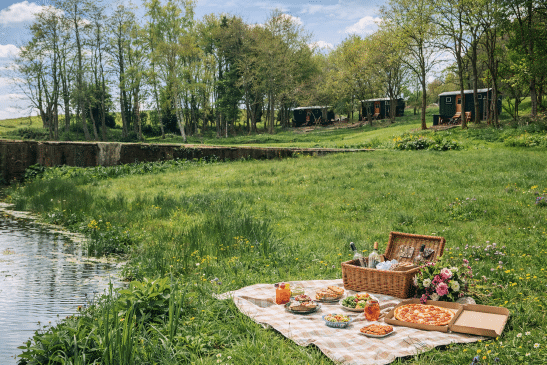 A large picnic spread with pizza, salads, a wicker hamper, and flowers on a checkered blanket in a grassy field near a lake, with three shepherd's huts in the background.
