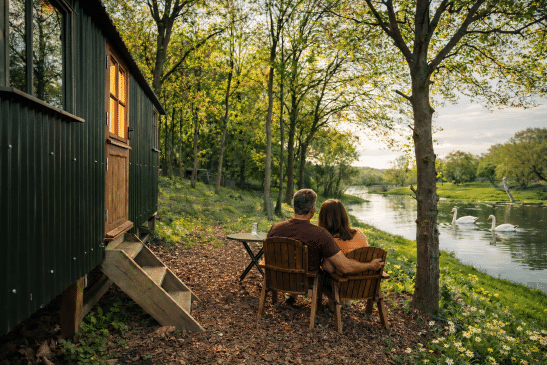 A man and woman sit closely in wooden armchairs on a woodchip clearing outside a dark green shepherd's hut with a glowing window, looking out at two swans swimming on a river during sunset.