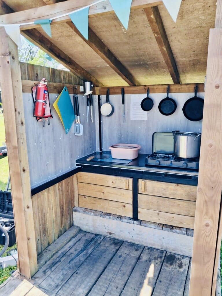 Covered outdoor kitchen at Glamp and Tipple with a camping stove, cast-iron pans hung on hooks, utensils and a prep counter under a bunting-strung timber roof