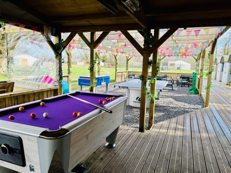 Covered games area at Glamp and Tipple with a purple-baize pool table, table football and table tennis under a bunting-decked pergola, seating and a yurt beyond