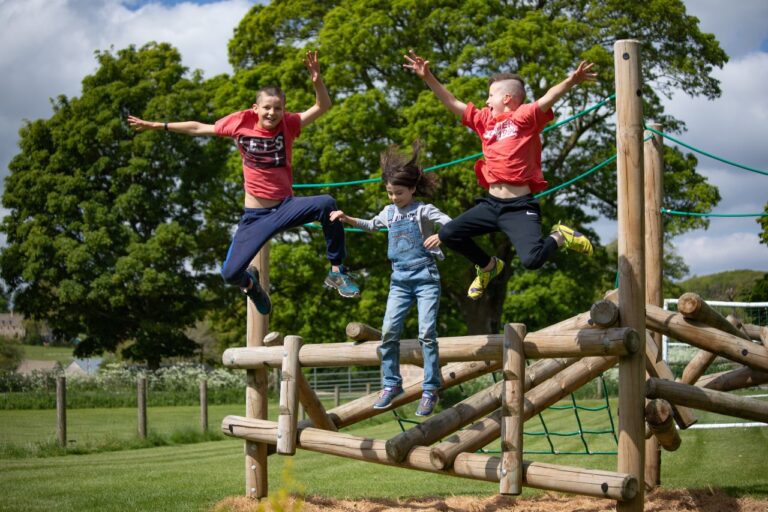 Three children leaping from a timber adventure play frame in a field, Notgrove Holidays, Cotswolds
