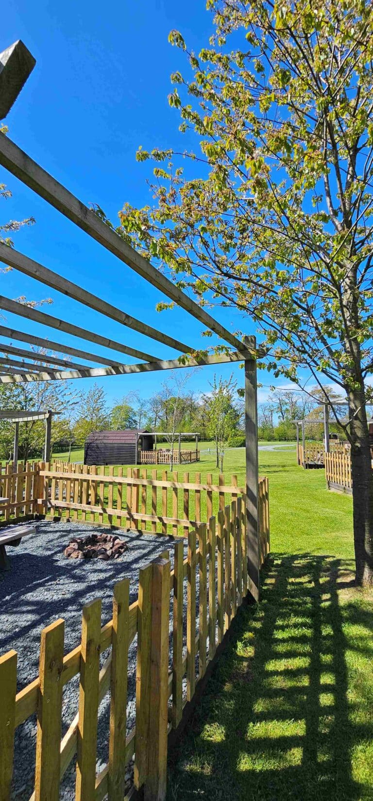 View across the glamping site of Meadow Falls, the sky is bright blue and the glamping pod has a gravel terrace with pickett fence around and stone fire pit with picnic bench