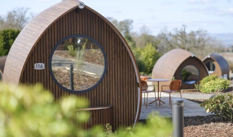 Sleek pointed-arch timber glamping pods with large round windows at Murrayshall Country Estate, one numbered 116, with cafe tables on a paved terrace and countryside views
