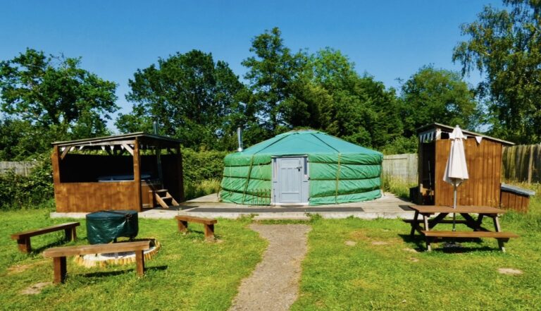 Green yurt on a timber deck at Glamp and Tipple, with a hot-tub shelter to one side and a covered kitchen cabin to the other, fire-pit benches on the lawn