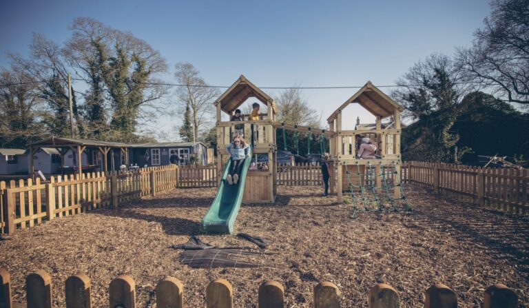 Children playing on the wooden climbing frame and green slide in the fenced bark-chip play area at Glamp and Tipple