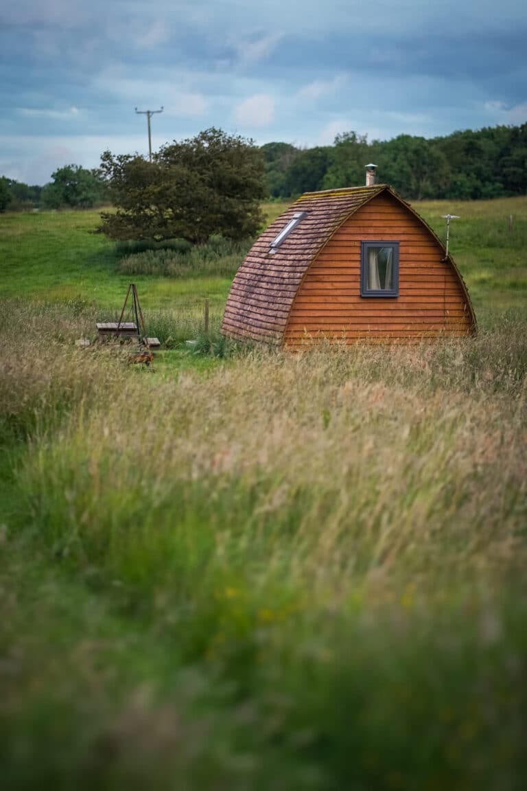 A cedar camping pod with a shingled curved roof and flue in a meadow of long grass at dusk at Tranwell Farm