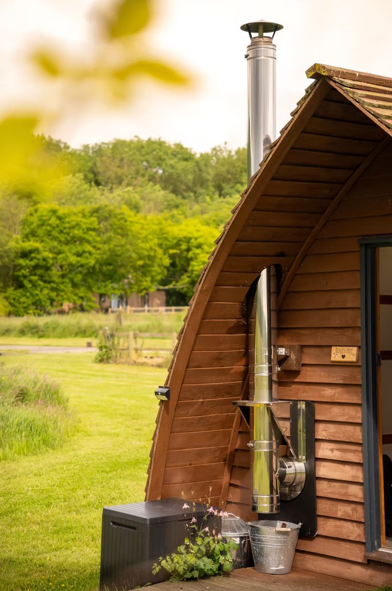 Exterior of the Rosie's Hut camping pod at Tranwell Farm with a shingled arched roof, wood-burner flue, a storage box and flowers on the decking