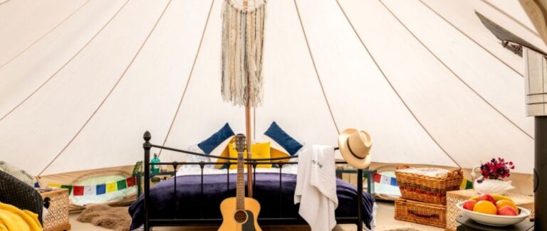 Inside the furnished bell tent with a black iron double bed, a macramé dreamcatcher on the centre pole, navy and yellow cushions, a guitar, wicker hampers and a bowl of fruit