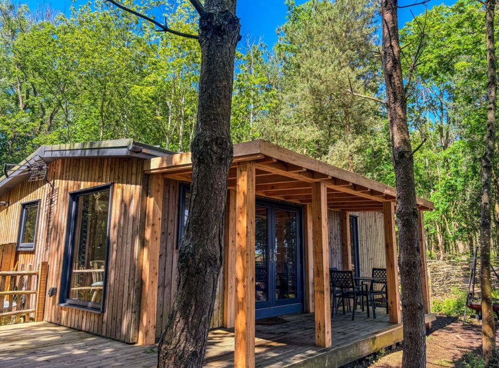 Timber woodland lodge at Tranwell Farm with a covered decked veranda and bi-fold doors, set among pine and birch trees