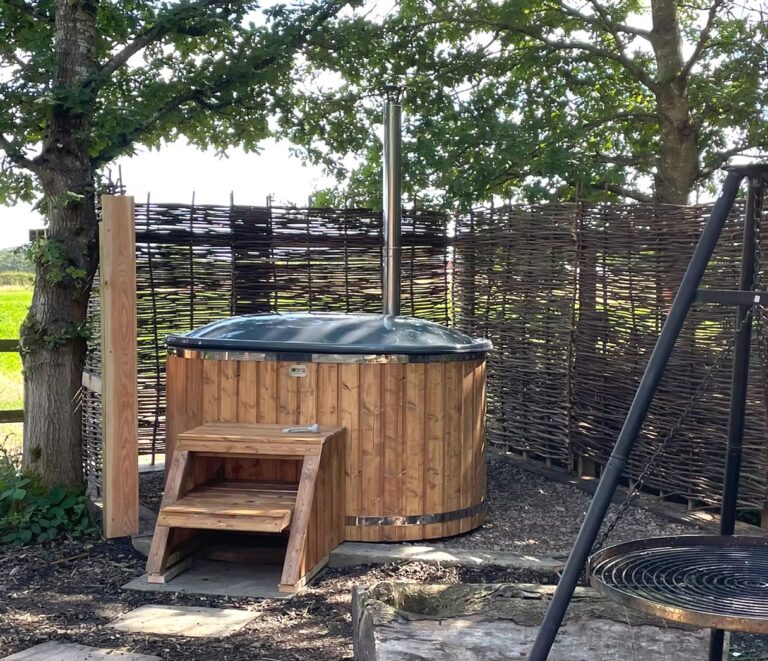Wood-fired hot tub with a cover, wooden steps and a willow privacy screen beside a swing fire pit under an oak tree at Tranwell Farm