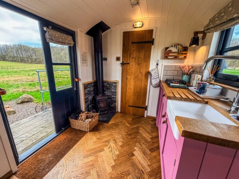 Inside the Poppy shepherd's hut at Tranwell Farm with a pink kitchen and Belfast sink, a wood-burning stove and a herringbone floor, the stable door open to the field