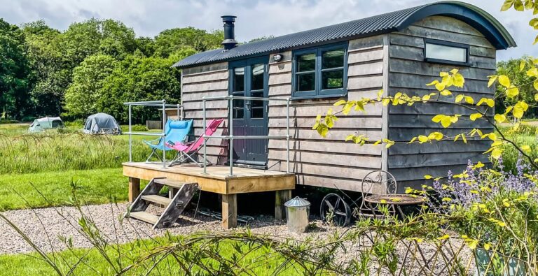 Timber shepherd's hut with a curved corrugated roof and decked veranda at Tranwell Farm, with pink and blue deck chairs, a fire pit and bell tents in the meadow behind