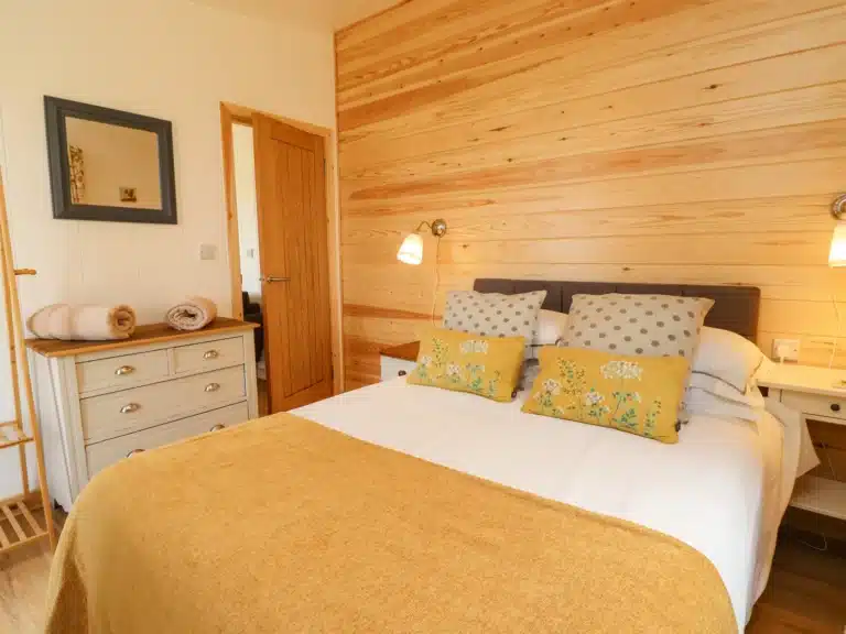 Pine-clad lodge bedroom at Tranwell Farm with a double bed, mustard throw and floral cushions, a painted chest of drawers, mirror and towels