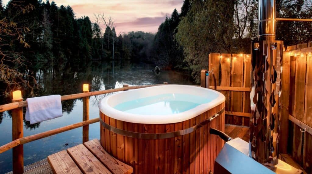 Wood-fired hot tub on a deck overlooking a lake at dusk, The Shepherds Hut Retreat Somerset