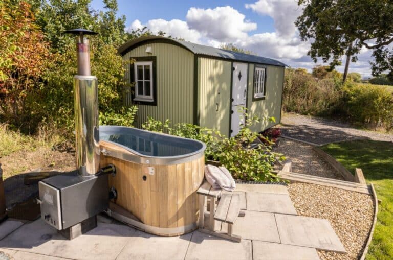 Green shepherd's hut with a wood-fired hot tub on the paved patio at Abbey Farm